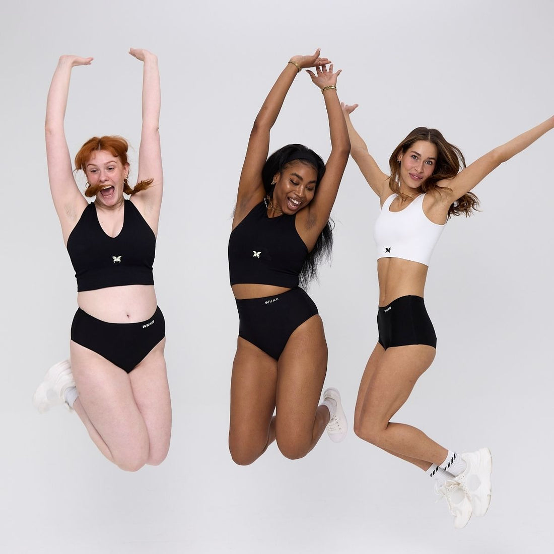 Three women jump joyfully in WUKA period underwear and crop tops, celebrating body confidence, movement, and menstrual empowerment in a bright studio setting.